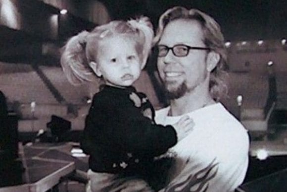 Black and white photo of James Hetfield in a t-shirt holding his daughter in a black dress.